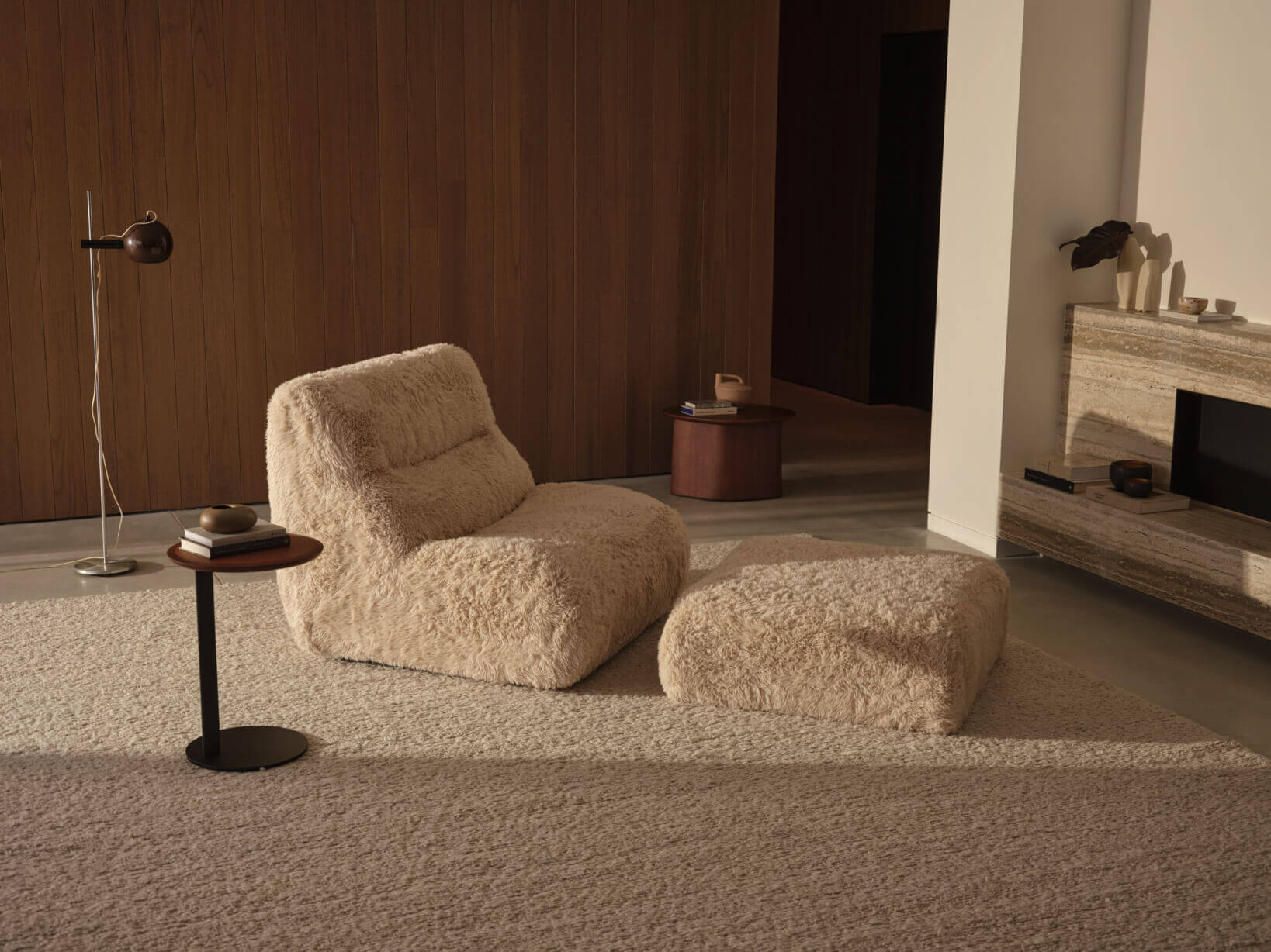 A matching high back sofa chair and ottoman, a perfect reading chair design, sit centrally in a contemporary living room, styled in a faux fur fabric on a soft, warm-toned rug.