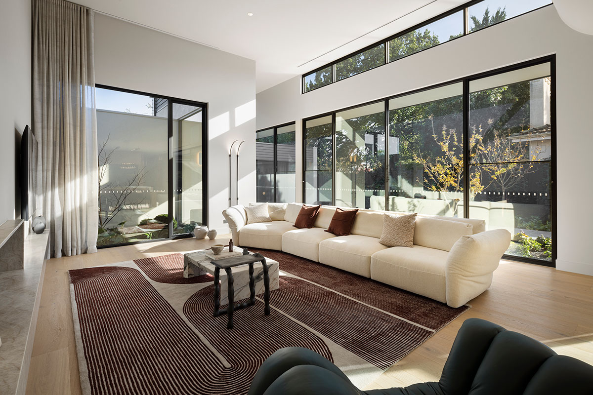 A modern living room with floor to ceiling windows and natural light. A beige modular sofa is decorated with brown cushions.