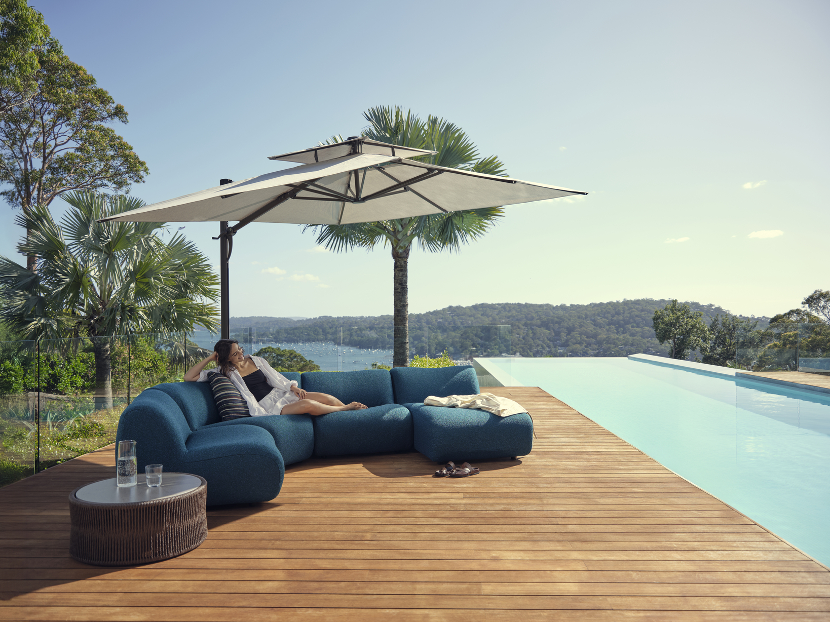 A woman lounges on a modular outdoor sofa styled in a blue bouclé fabric, the setting is shaded by an outdoor umbrella and overlooks a pool and landscapes.